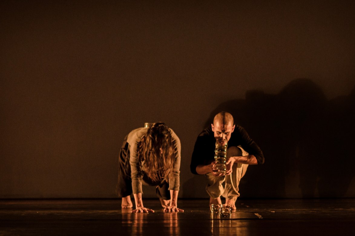 Foto de una pareja de bailarines en posición agachada sobre el piso, uno de los bailarines sopla dentro d eun tubo de vidrio.